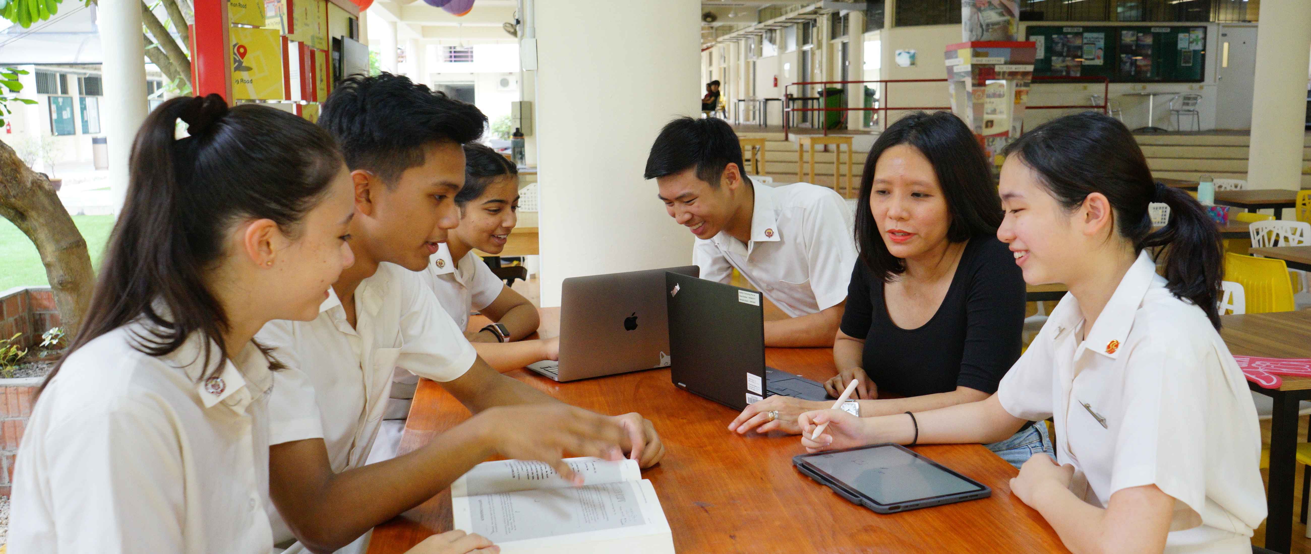 Five students in white uniforms and a woman in black sit at a wooden table with laptops and a tablet, studying together.